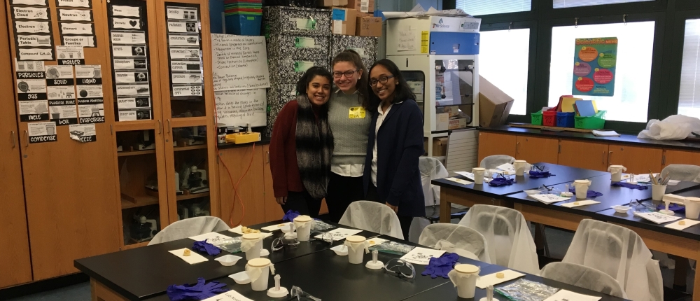 Three students smiling together behind desks with hands-on science projects on them